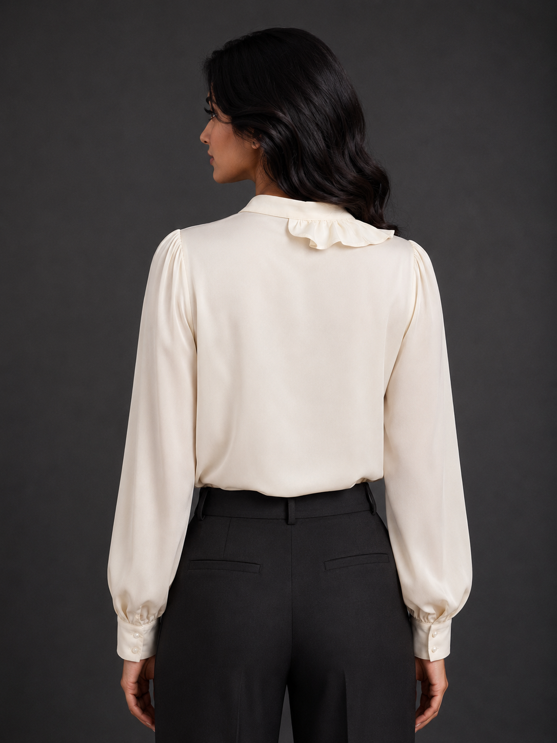 Woman wearing a cream blouse with long sleeves and black pants against a gray background