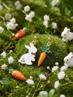Pair of bunny and carrot earrings on a green moss background with small white flowers and carrots.