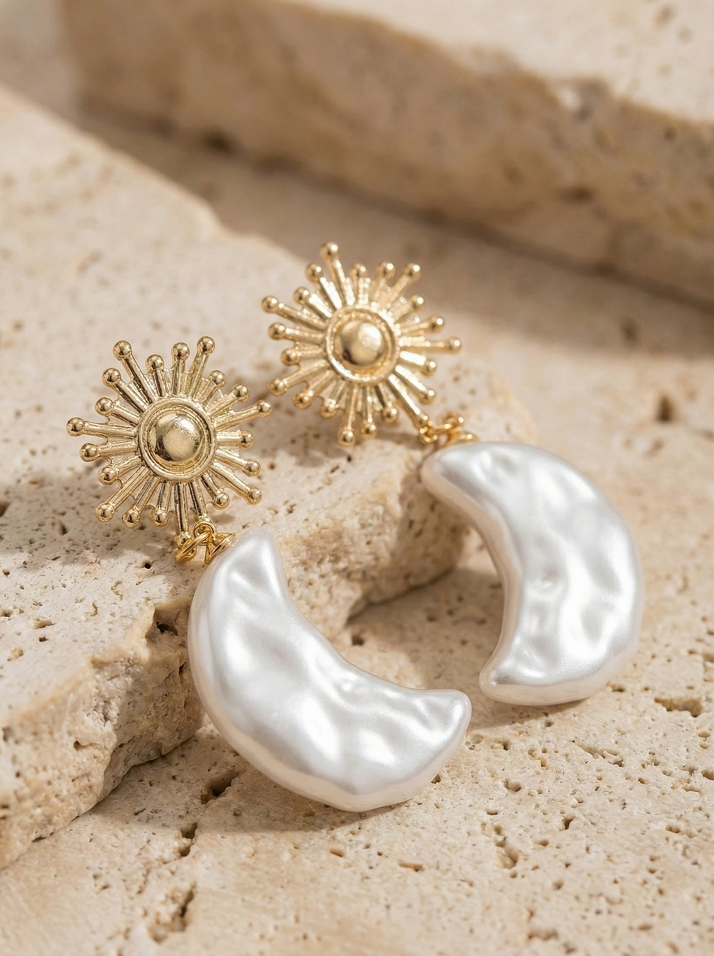 Gold sunburst earrings and a white crescent moon earring on beige stone surface