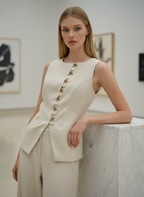 Woman wearing a cream-colored sleeveless blazer with gold buttons in an art gallery setting.