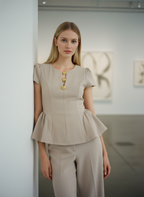 Woman wearing a beige peplum top with gold buttons and matching pants in an indoor setting.