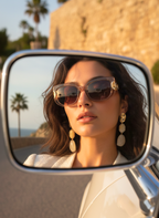 Woman wearing sunglasses and earrings reflected in a car mirror with palm trees and a building in the background.