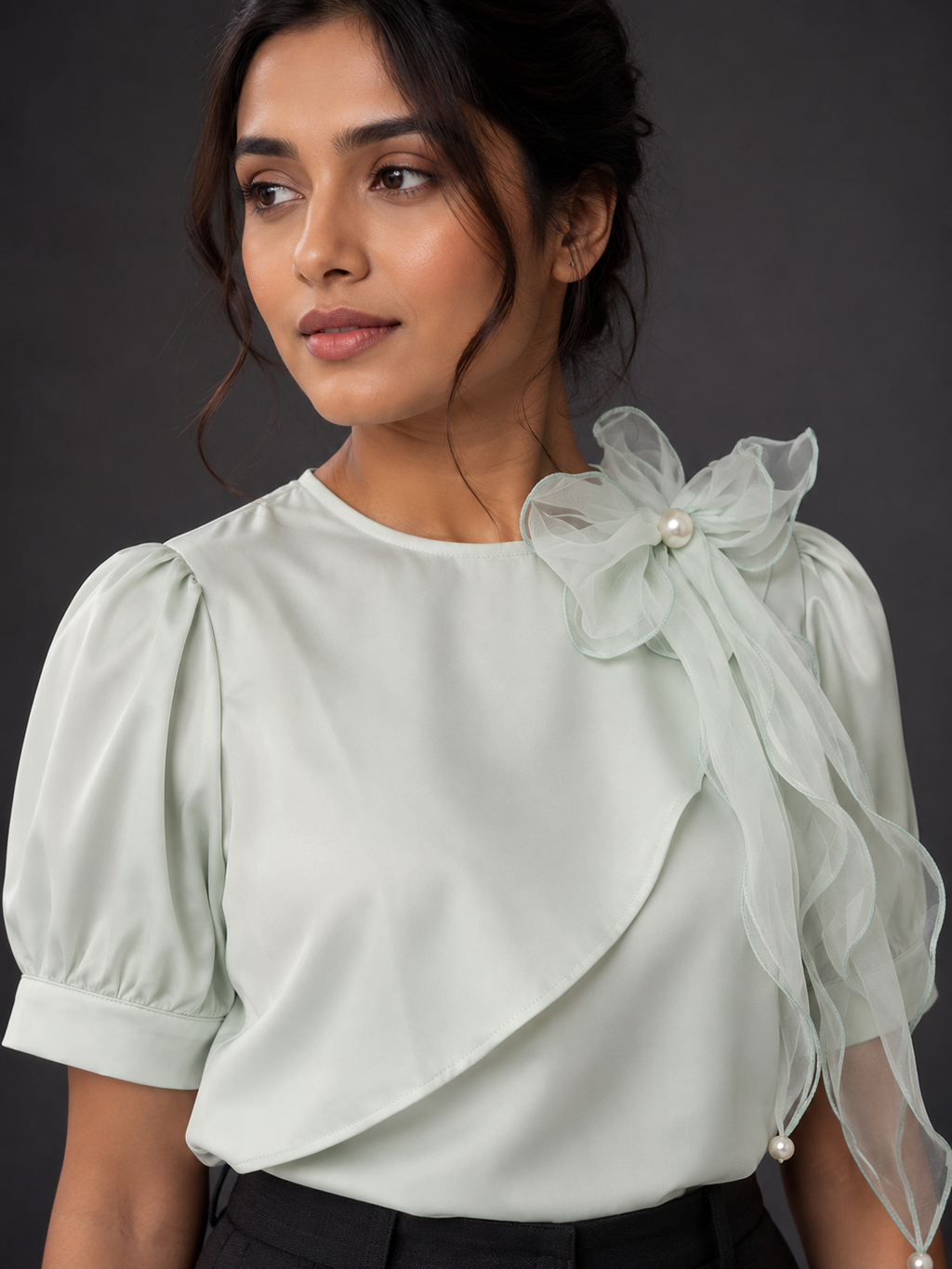 Woman wearing a light green blouse with sheer sleeves and a floral bow detail on a dark background